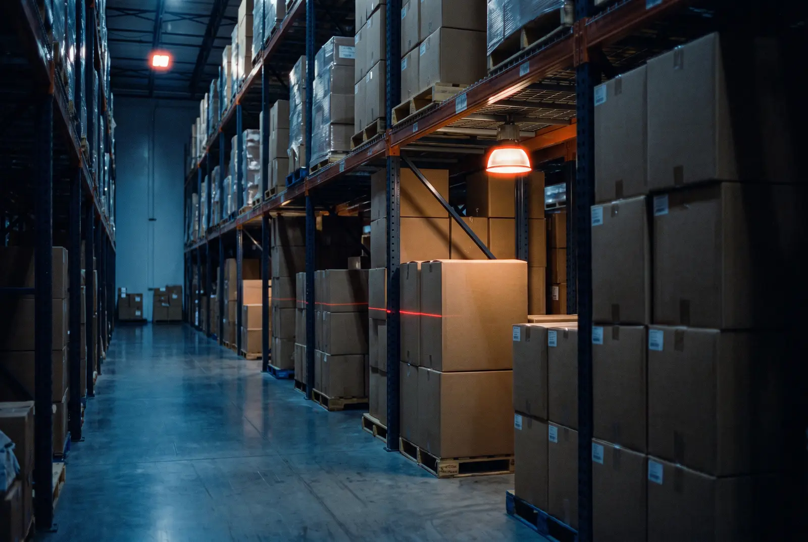 Dark warehouse aisle with tall pallet racking full of cardboard cartons and a red scanner beam crossing one of the boxes